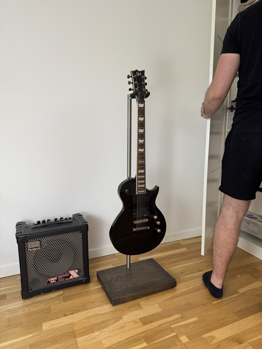 Dark electric guitar on a handcrafted stand with a solid wood base, displayed beside a guitar amplifier in a modern room.